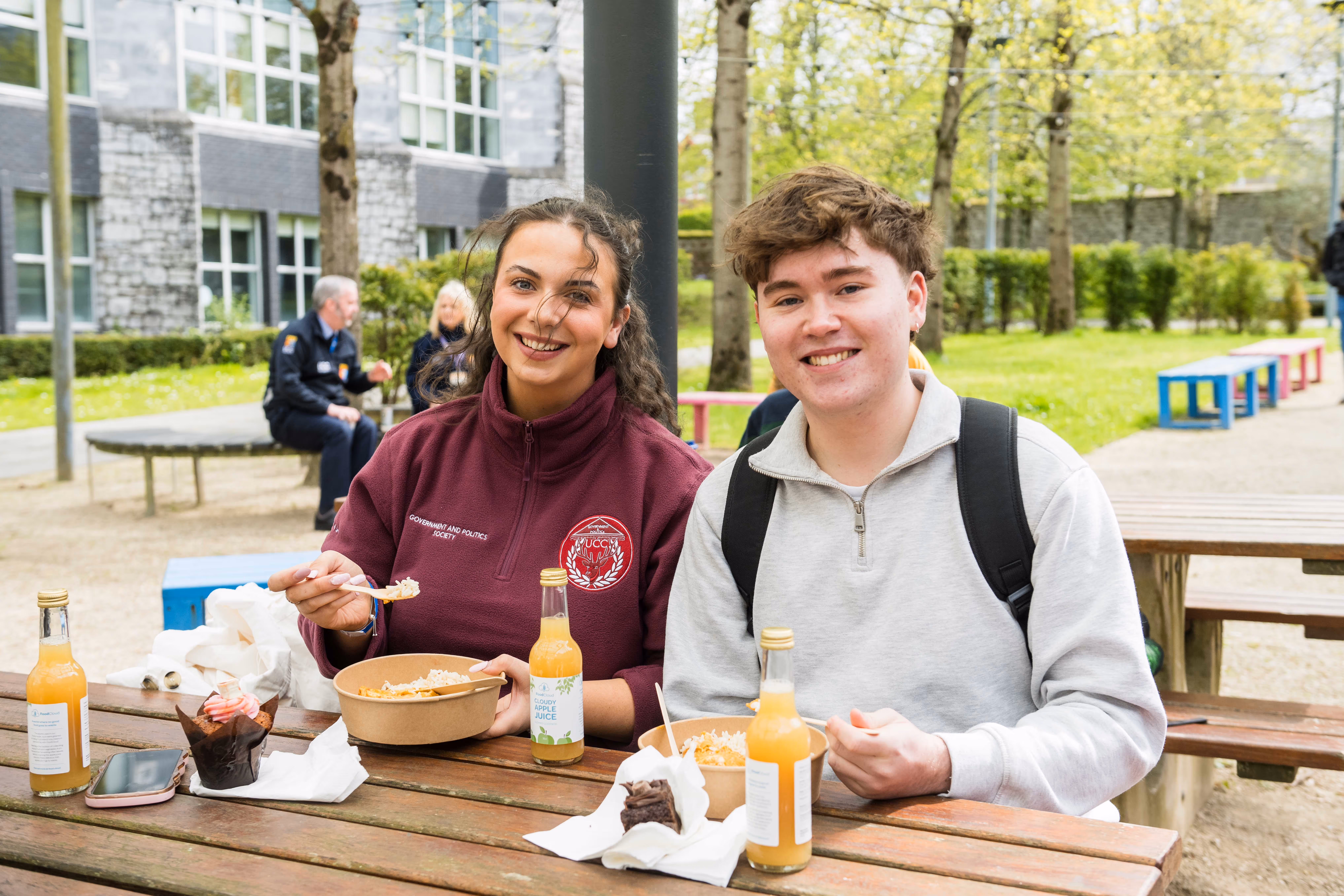 Two students sitting at a wooden picnic table outdoors eating rice bowls with bottles of cloudy apple juice and desserts in front of them.