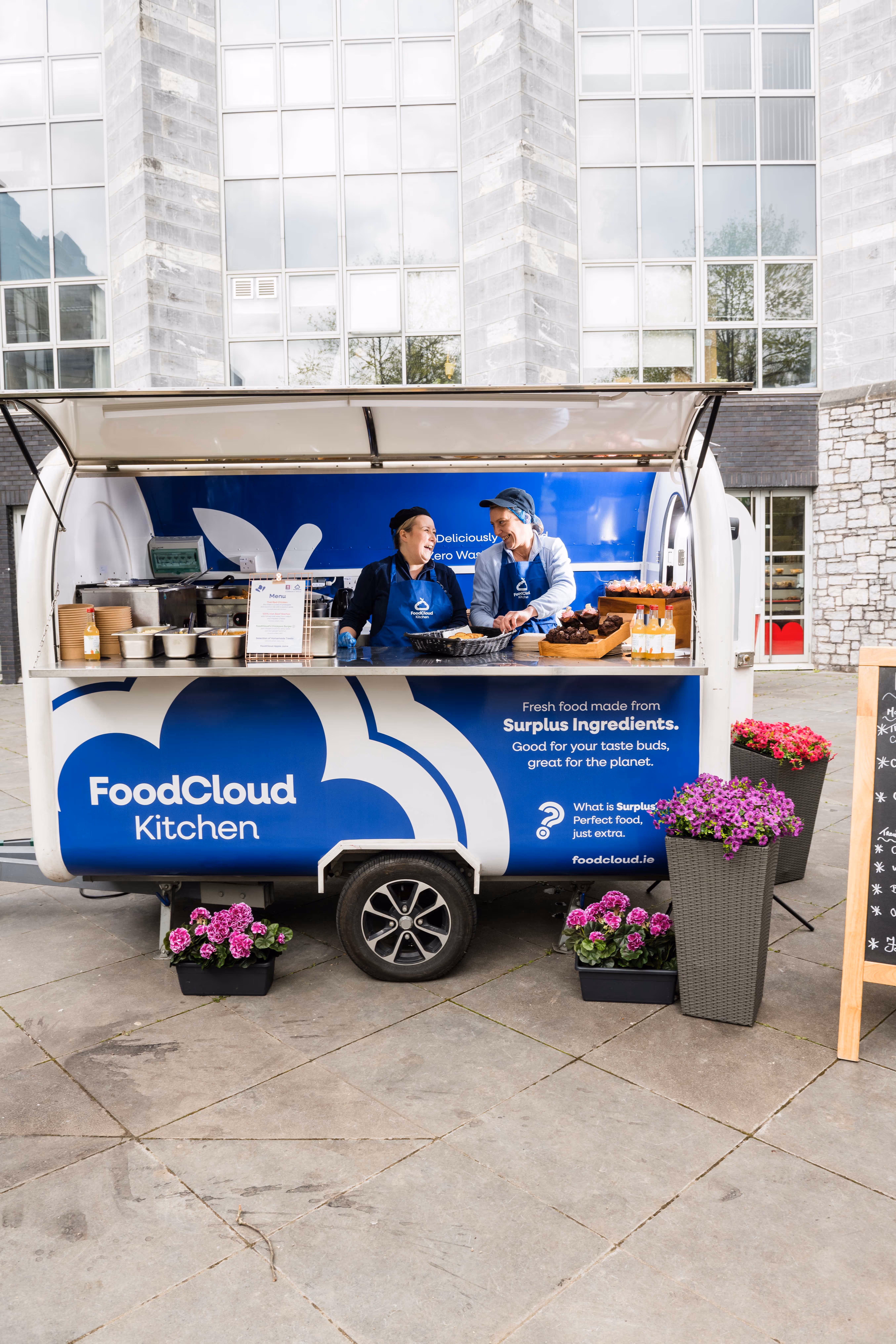 Two smiling people in aprons serving food from a FoodCloud Kitchen mobile food truck decorated with purple flowers.
