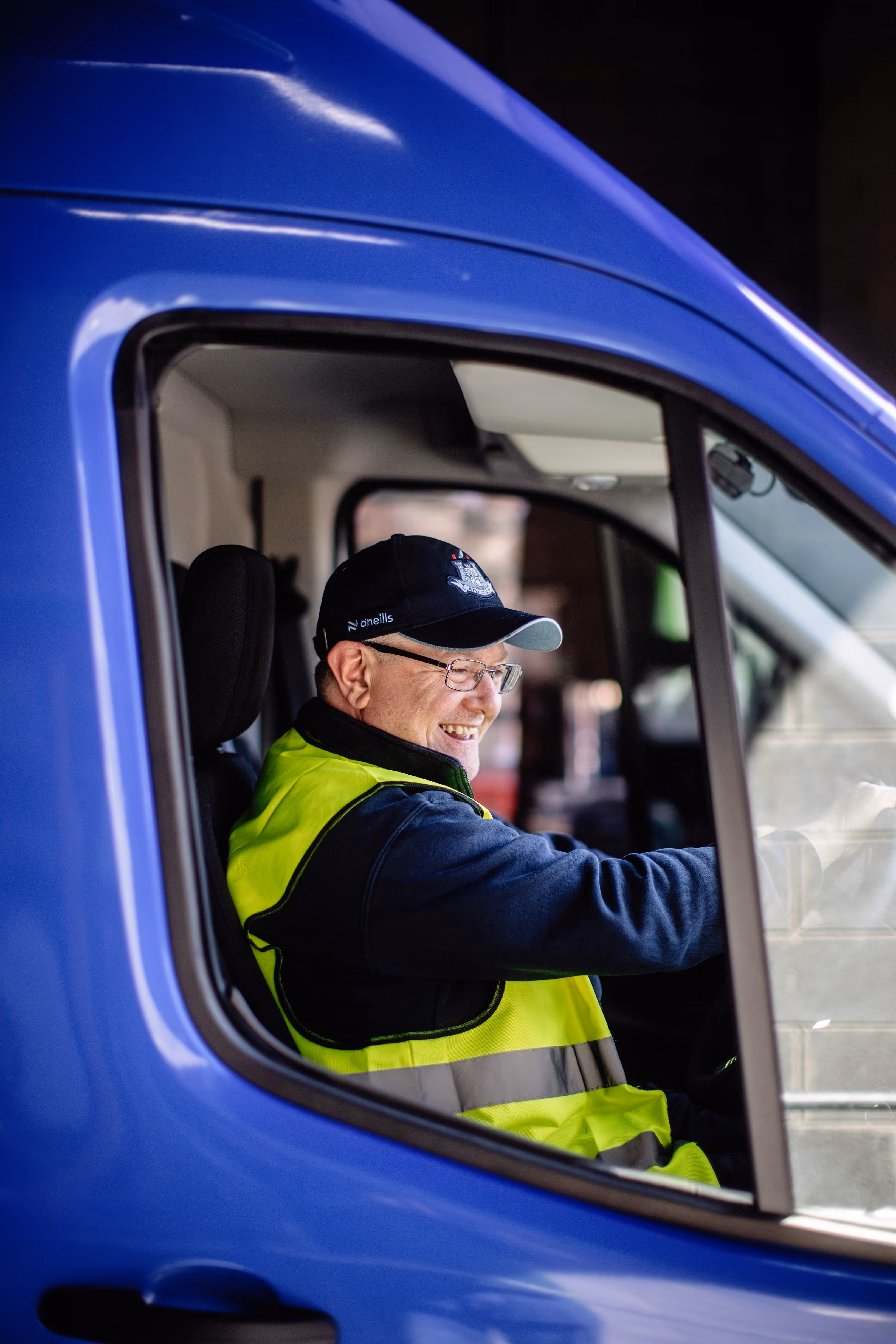 Smiling man wearing glasses, a navy cap, and a yellow safety vest driving a blue vehicle.