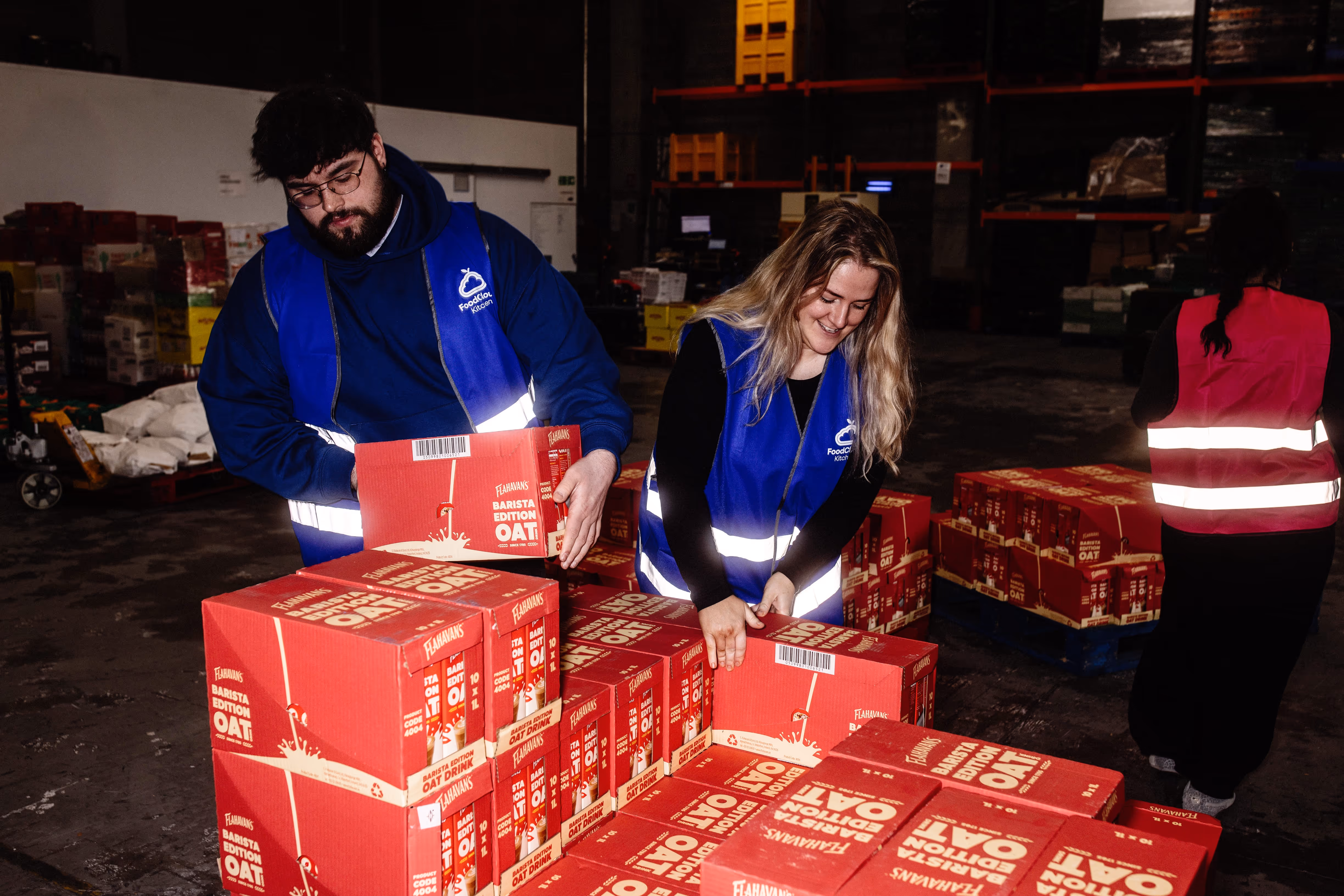 Two workers in blue vests handling stacked red boxes labeled Barista Edition Oat in a warehouse.