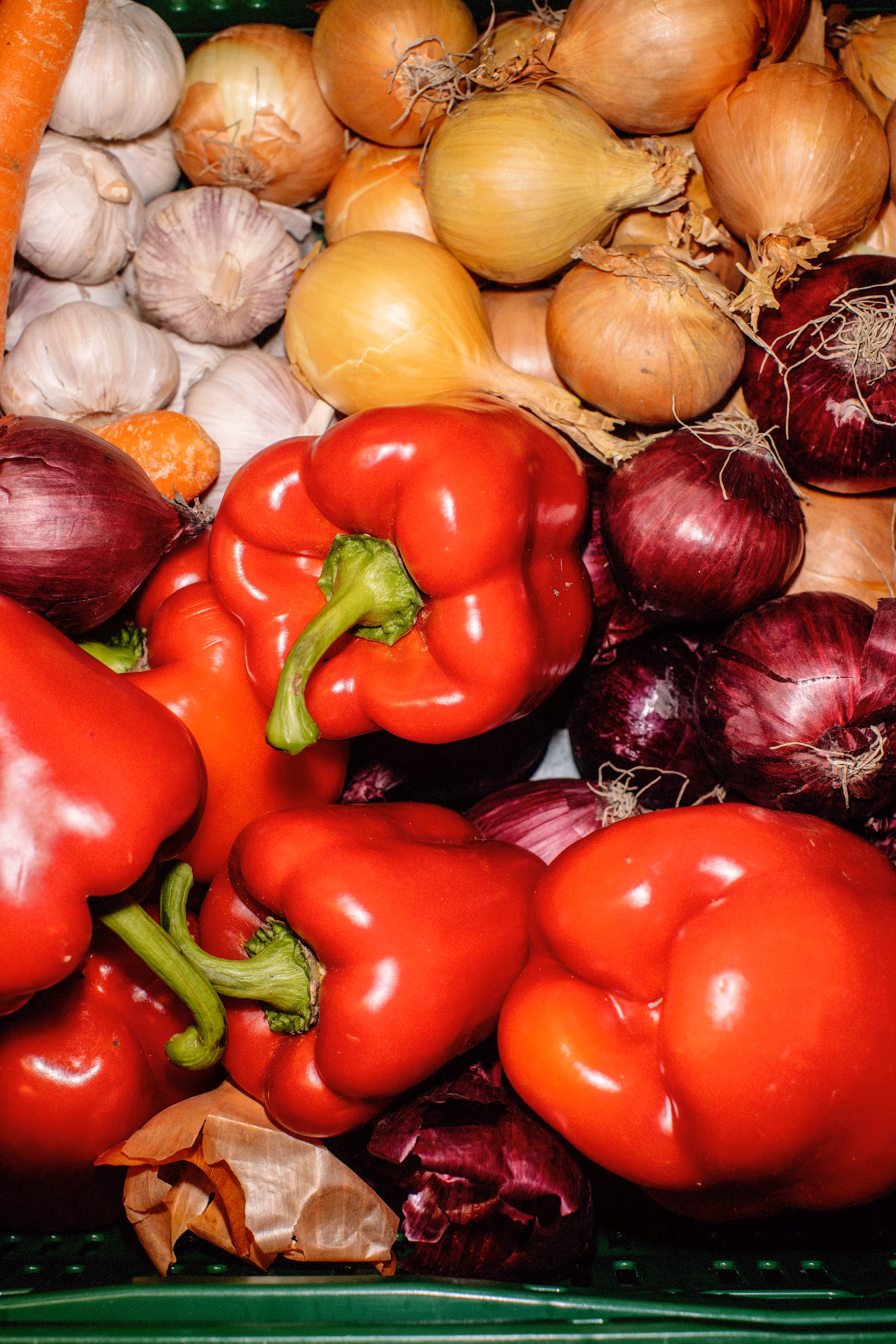 Close-up of red bell peppers, yellow onions, red onions, garlic, and a carrot in a crate.