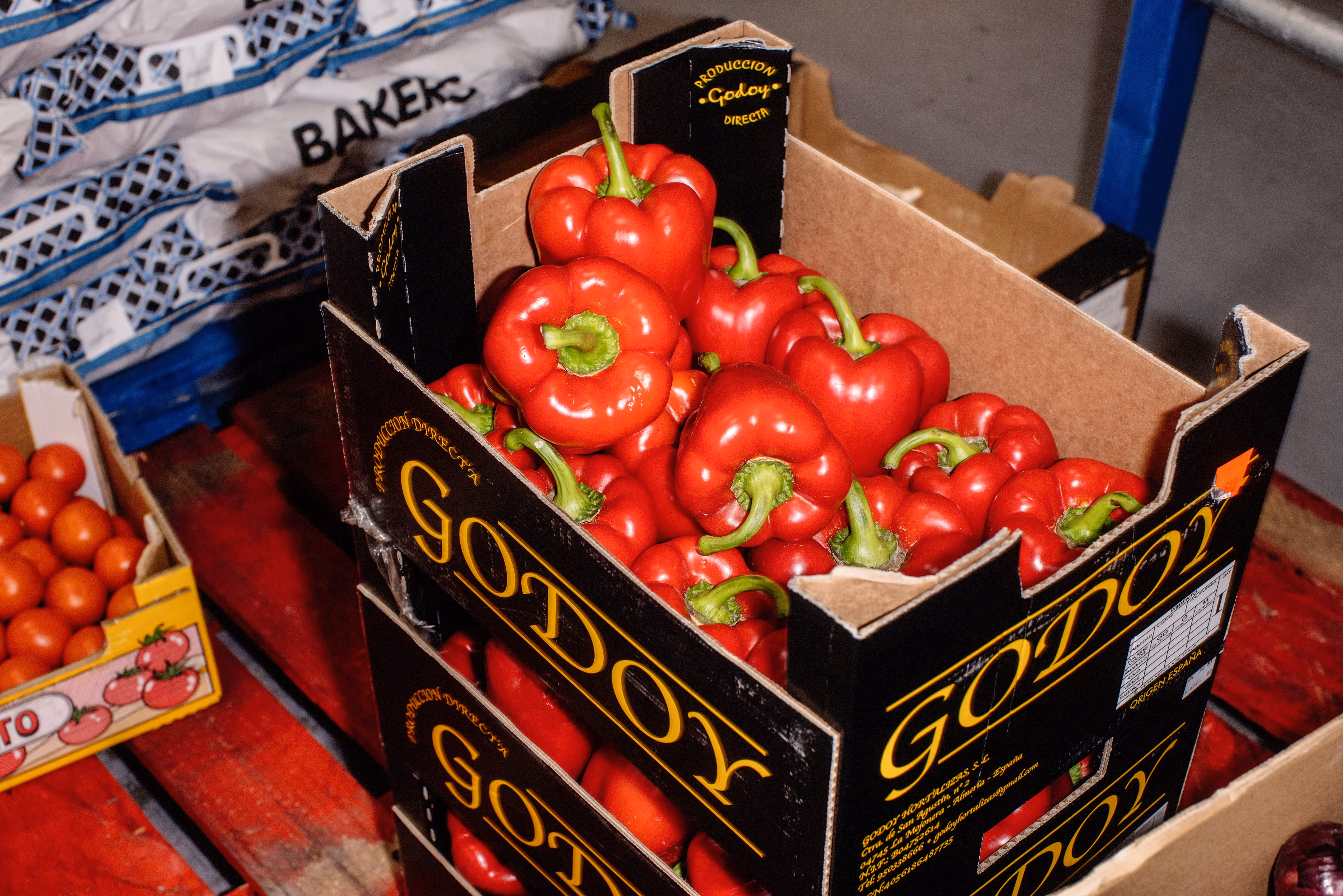 Box of fresh red bell peppers stacked on top of another box labeled 'Godoy' in a storage area.