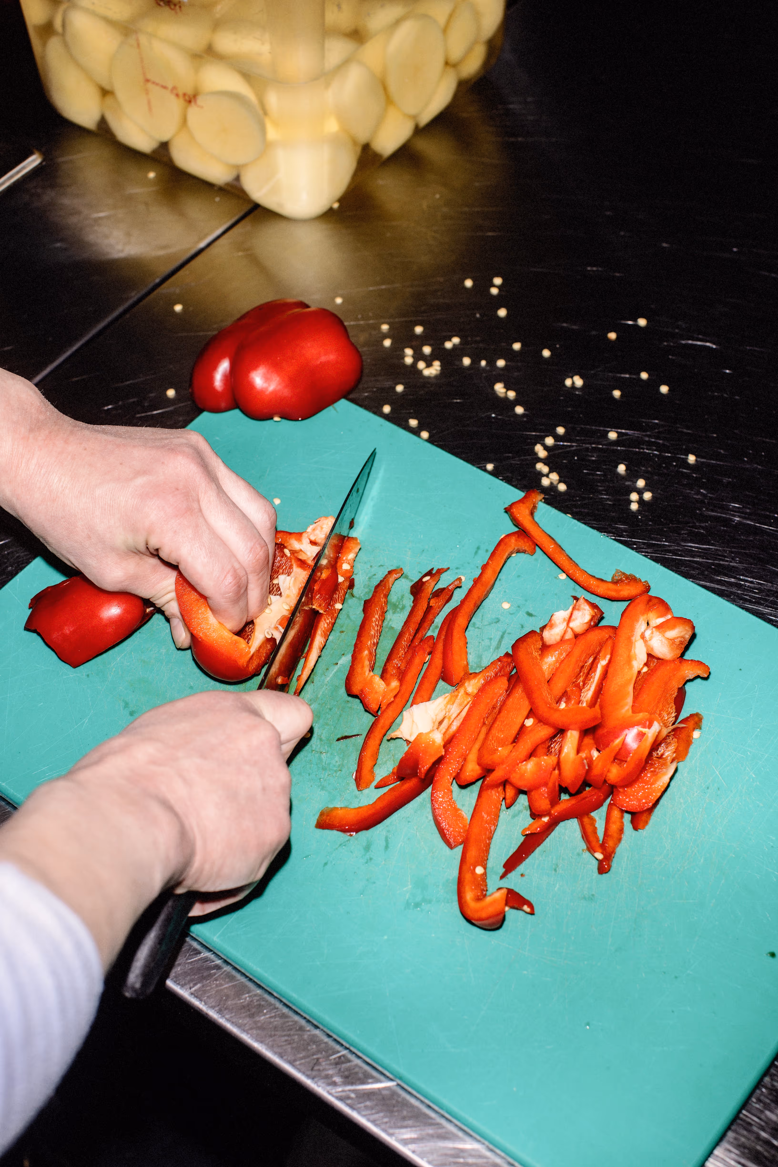 Hands slicing a red bell pepper into thin strips on a green cutting board with pepper seeds scattered around.