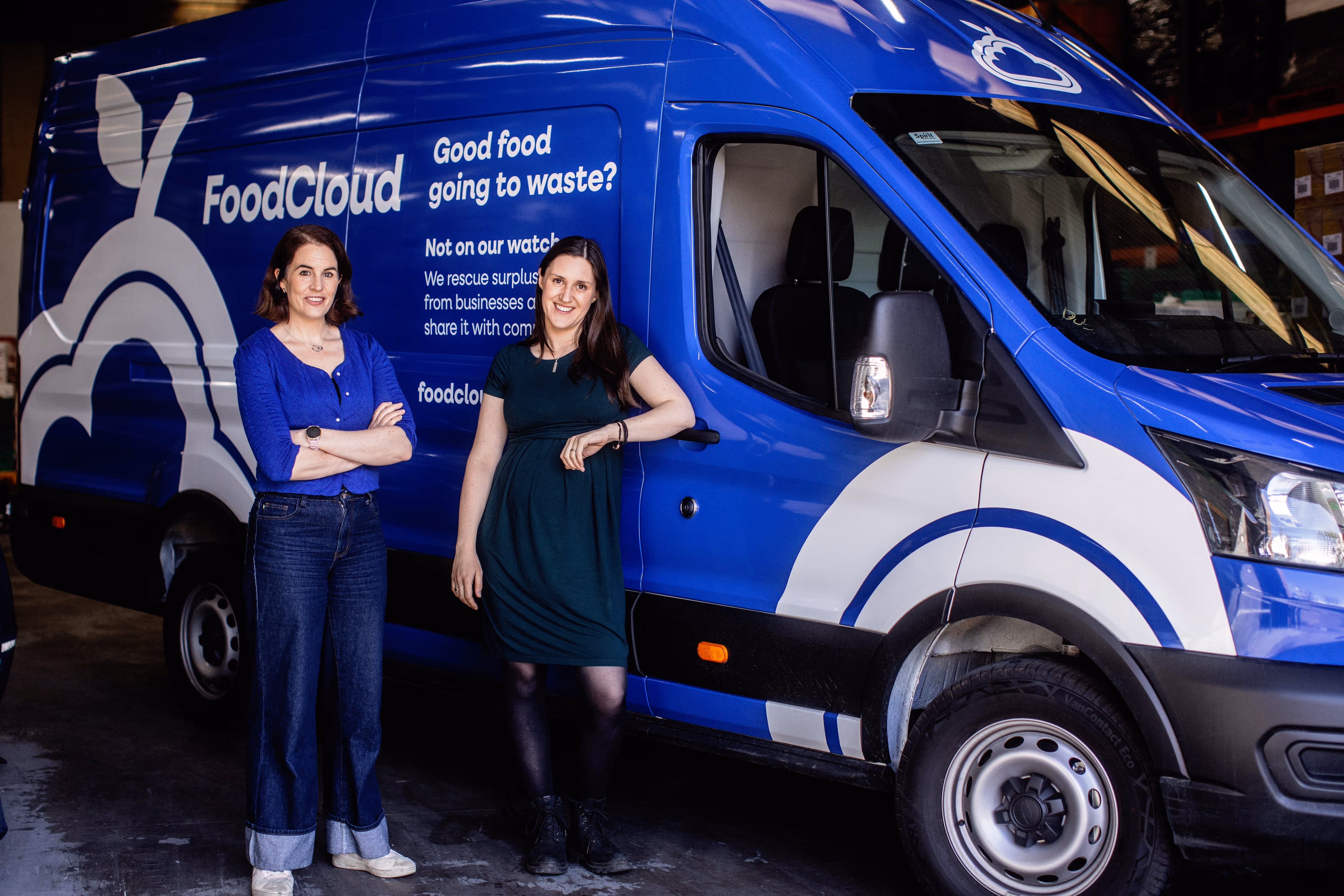 Two women standing and smiling in front of a blue FoodCloud delivery van with food rescue messaging.