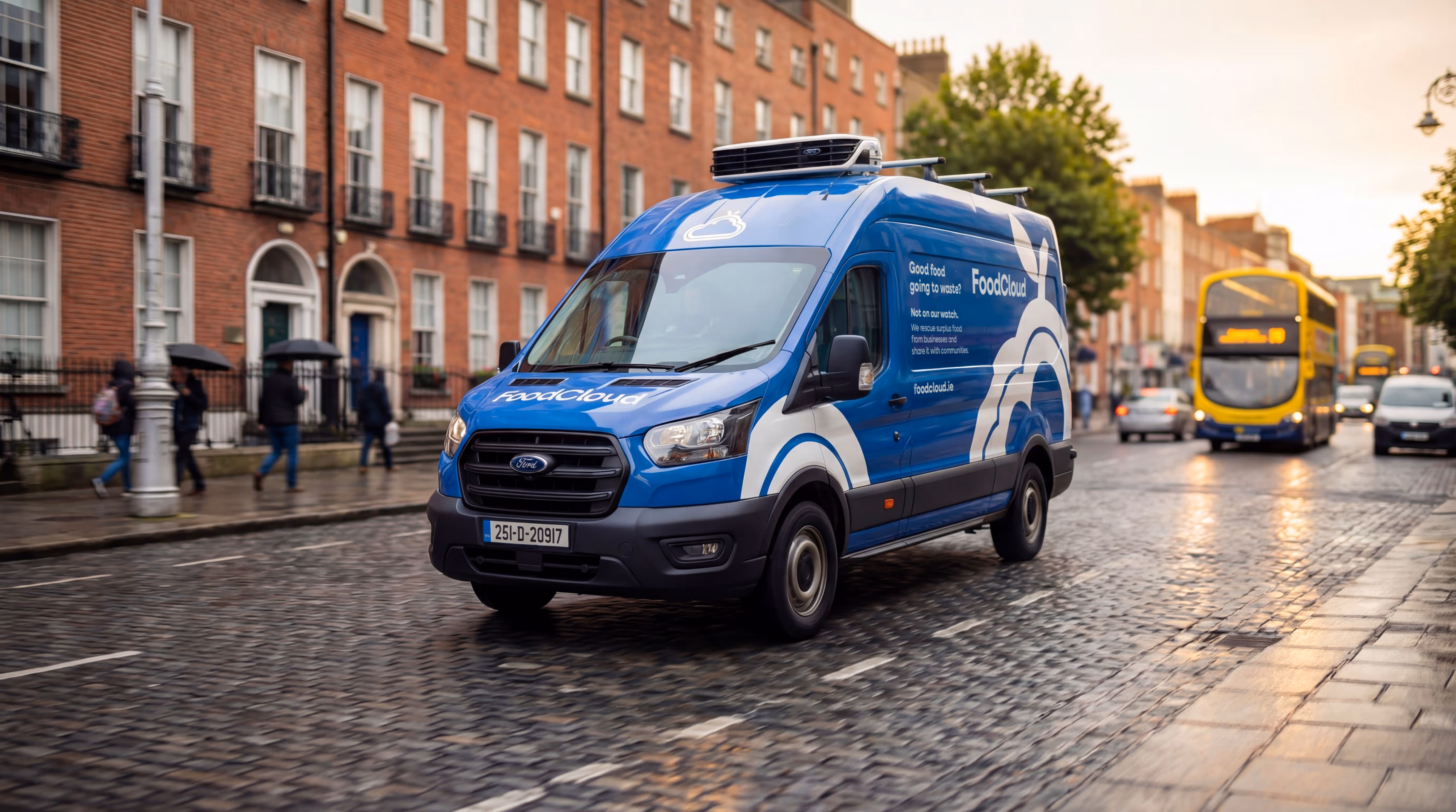 Blue FoodCloud van driving on a wet cobblestone street in a city with red brick buildings and a yellow double-decker bus in the background.