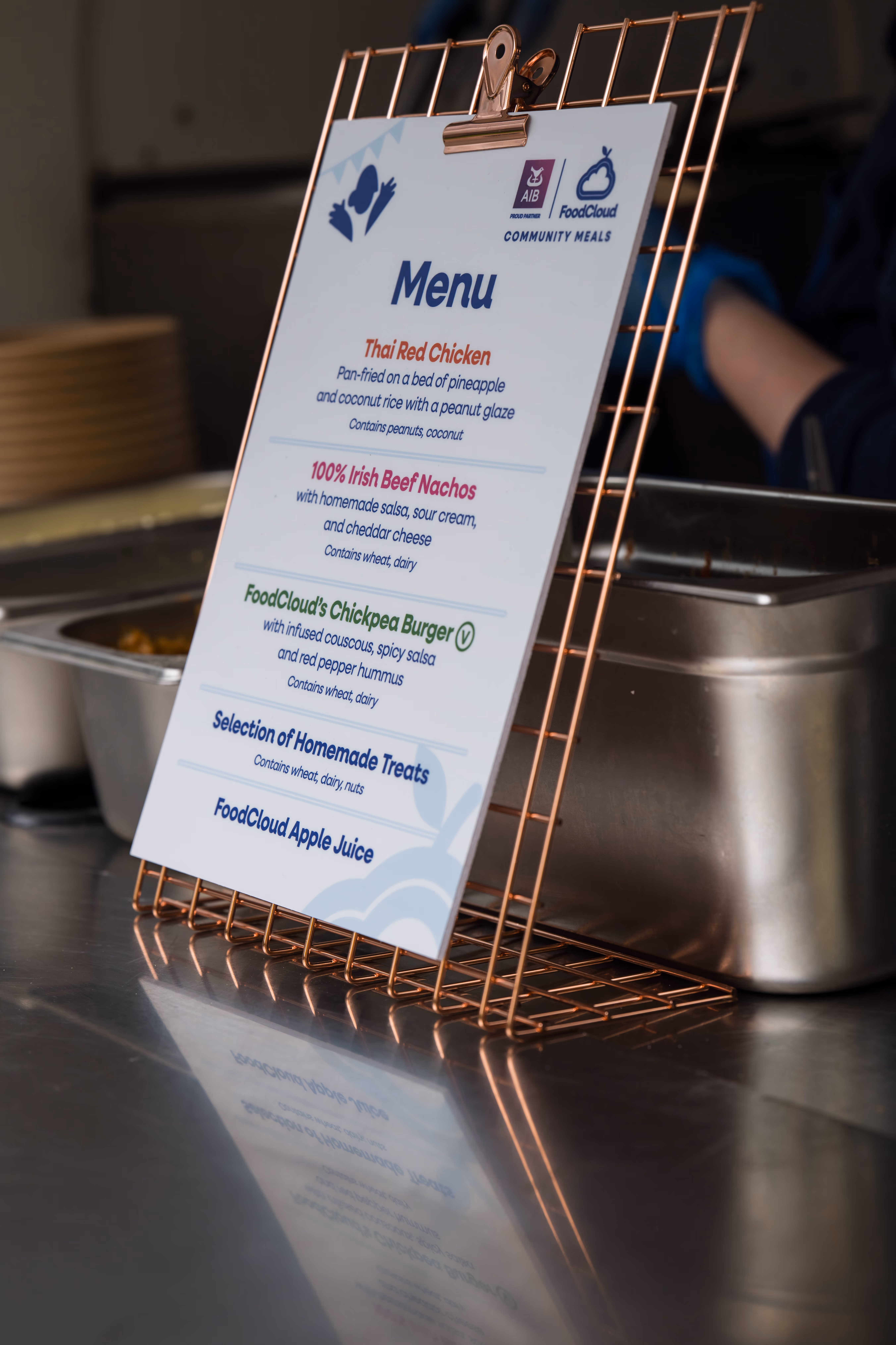 Menu on a wire stand listing Thai Red Chicken, 100% Irish Beef Nachos, FoodCloud's Chickpea Burger, homemade treats, and FoodCloud apple juice.
