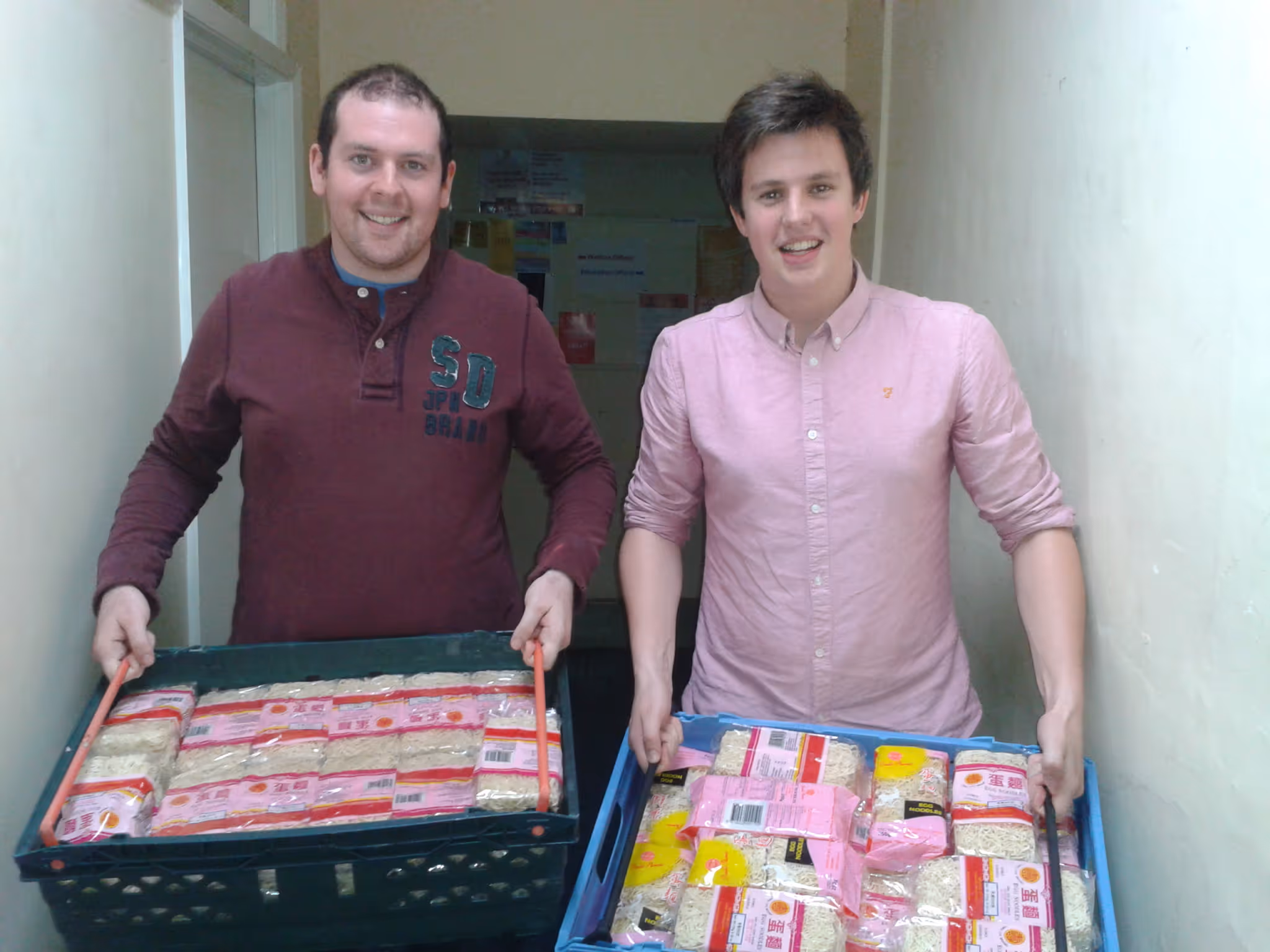 Two men smiling and holding crates filled with packaged instant noodles inside a hallway.