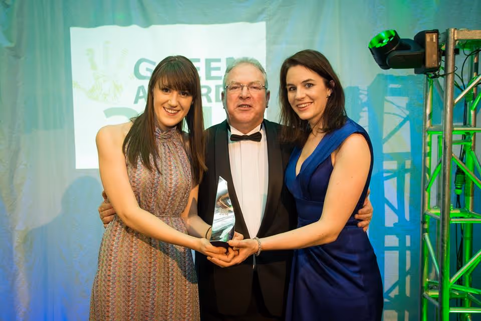 Three people dressed formally holding a triangular glass award at an event with a 'Green Awards' sign in the background.