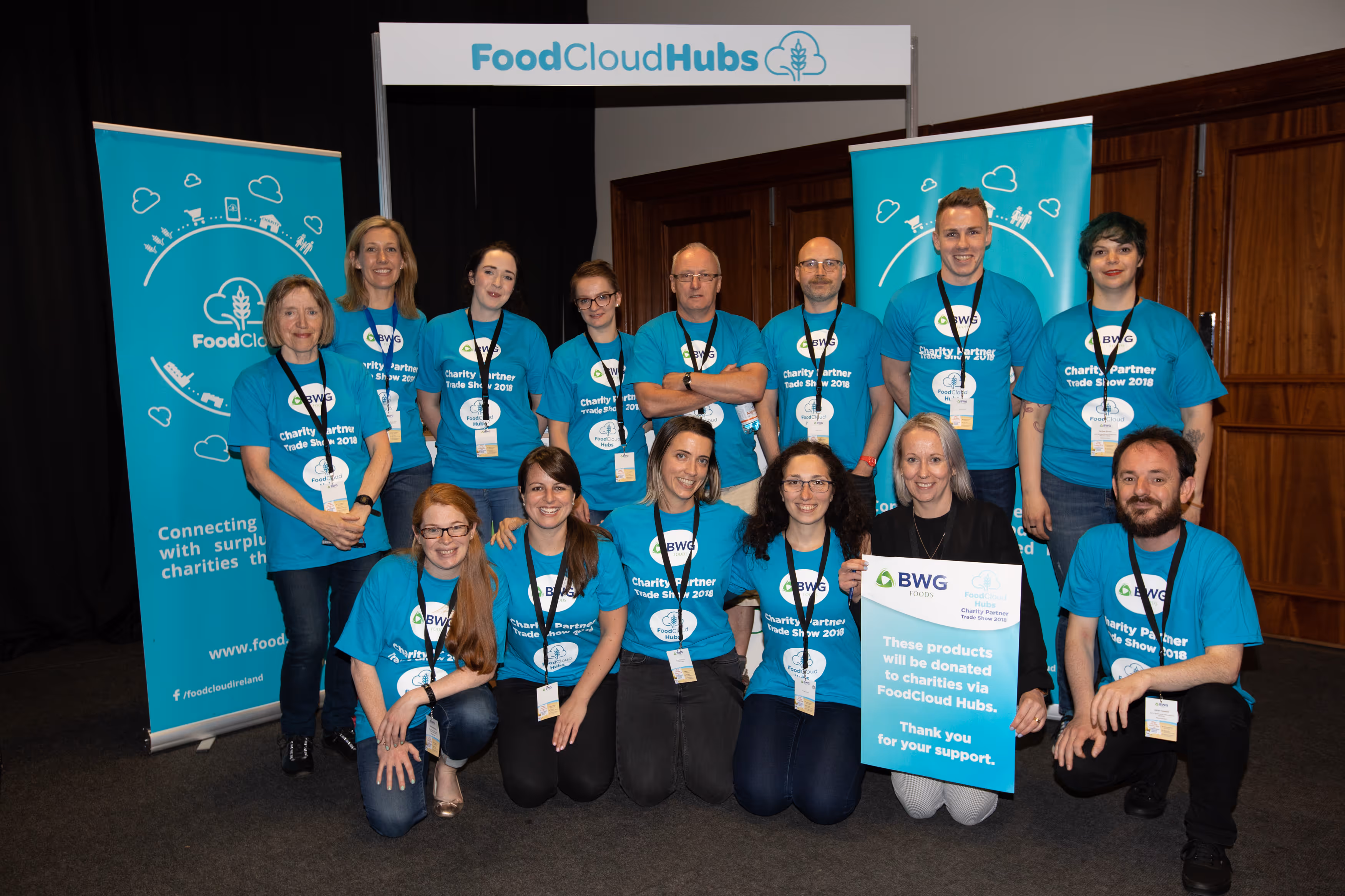 Group of 14 people wearing matching turquoise FoodCloud Hubs Charity Partner Trade Show 2018 t-shirts posing in front of FoodCloud Hubs signage and banners.
