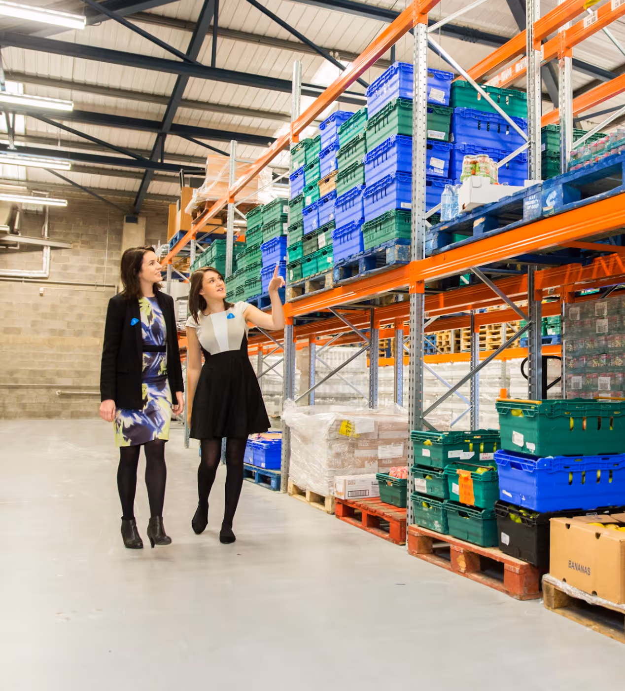 Two women walking and discussing in a warehouse aisle with shelves stacked with green and blue plastic crates and pallets.