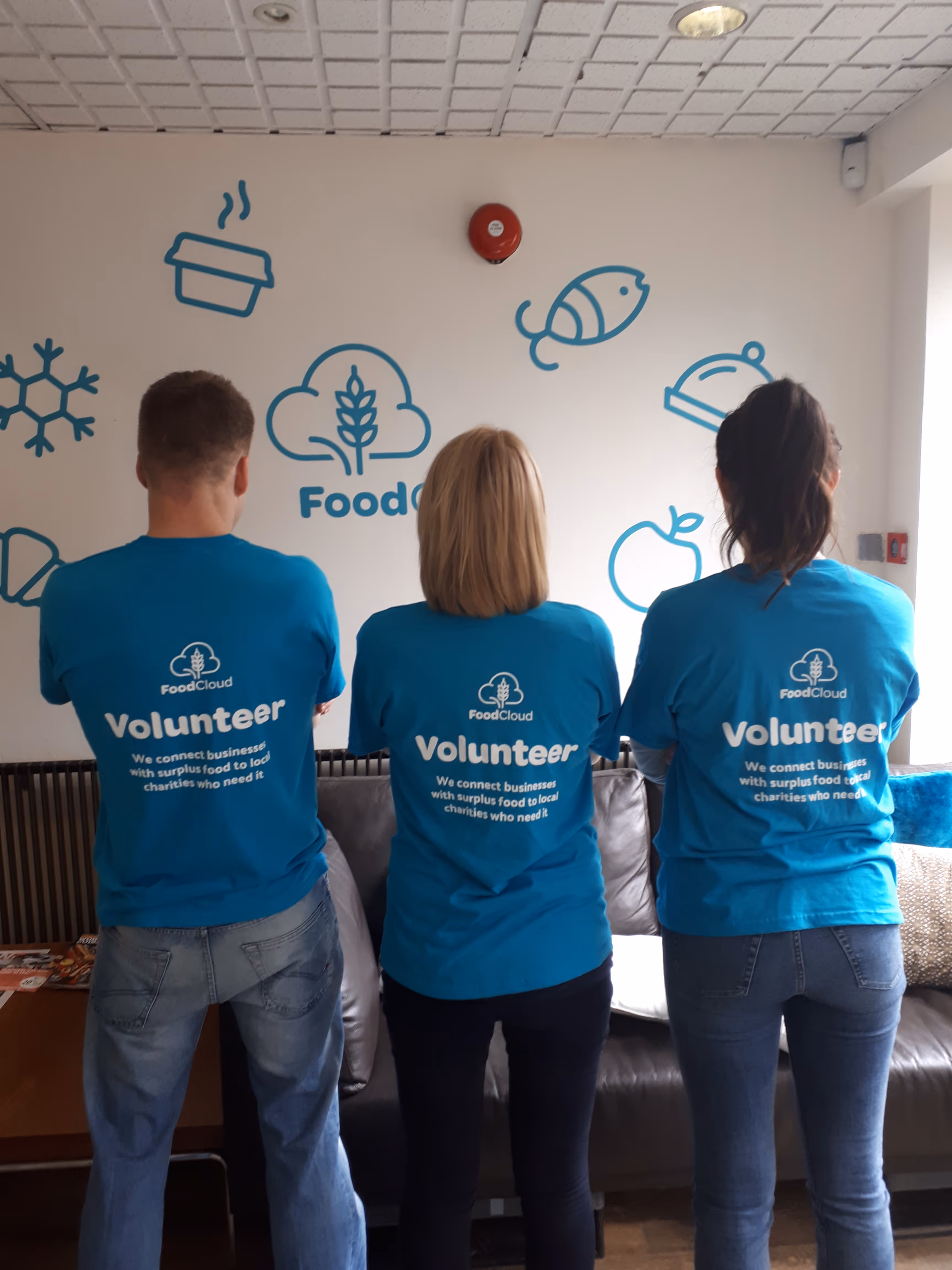 Three volunteers wearing blue FoodCloud shirts standing with their backs to the camera in front of a wall with food-related icons.