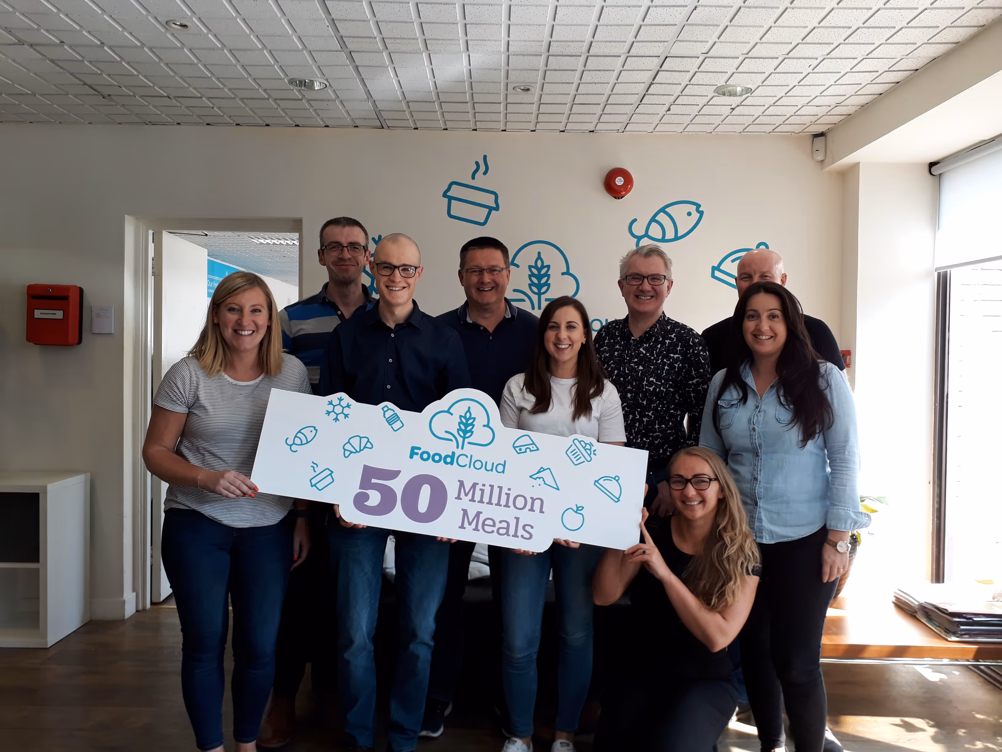 Group of nine people smiling indoors holding a sign reading 'FoodCloud 50 Million Meals' with food icons on it.