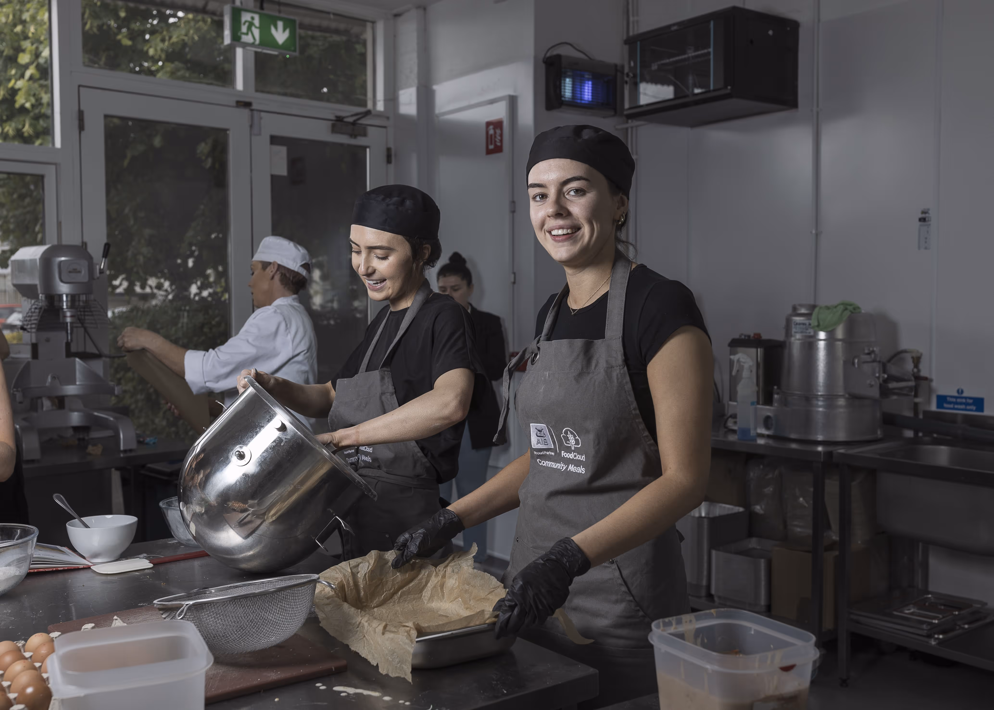 Two women wearing black hats and gray aprons preparing food in a commercial kitchen, one pouring from a large metal bowl and the other lining a baking pan with parchment paper.