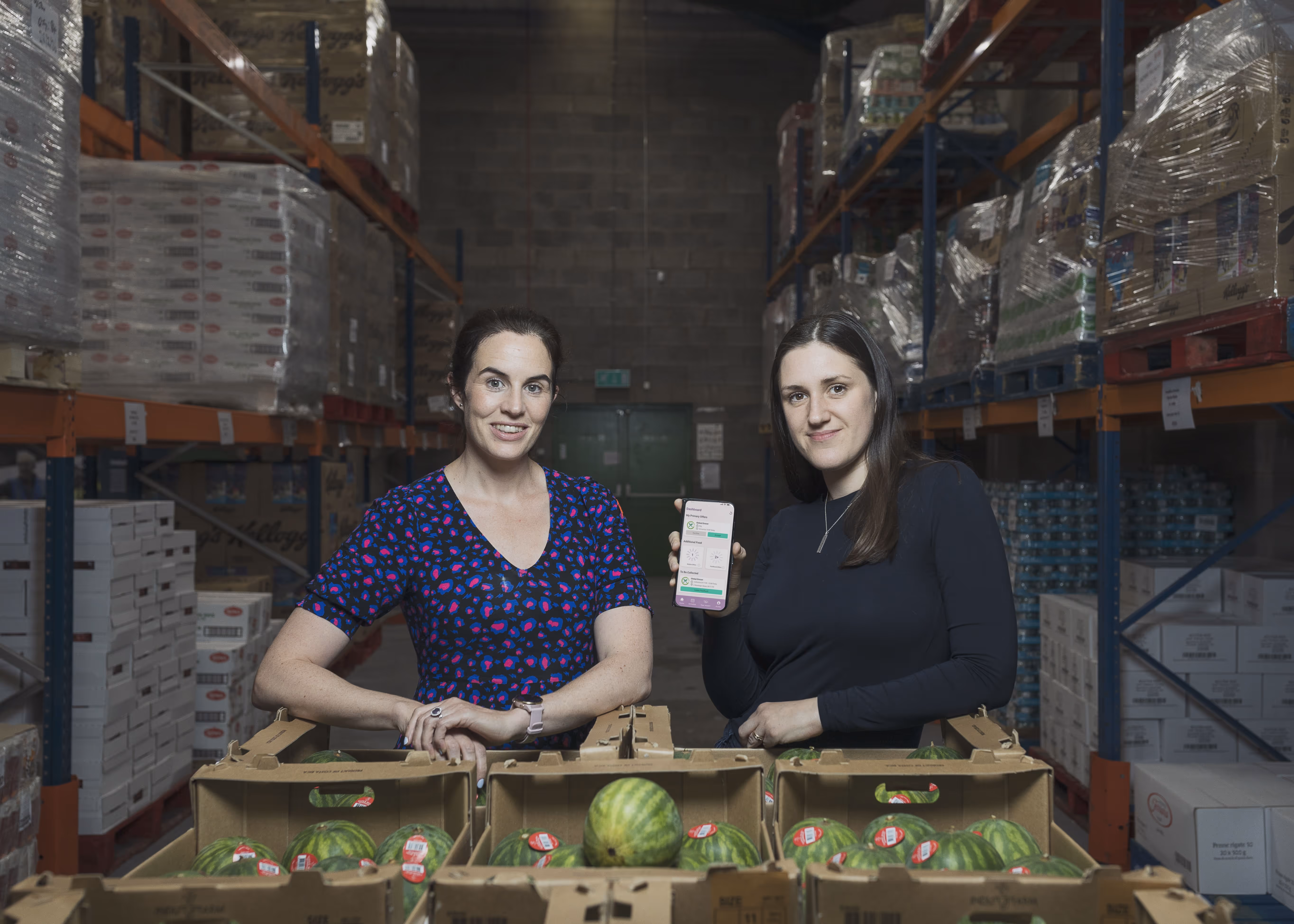 Two women standing behind boxes of watermelons in a warehouse, one holding a smartphone displaying an app.