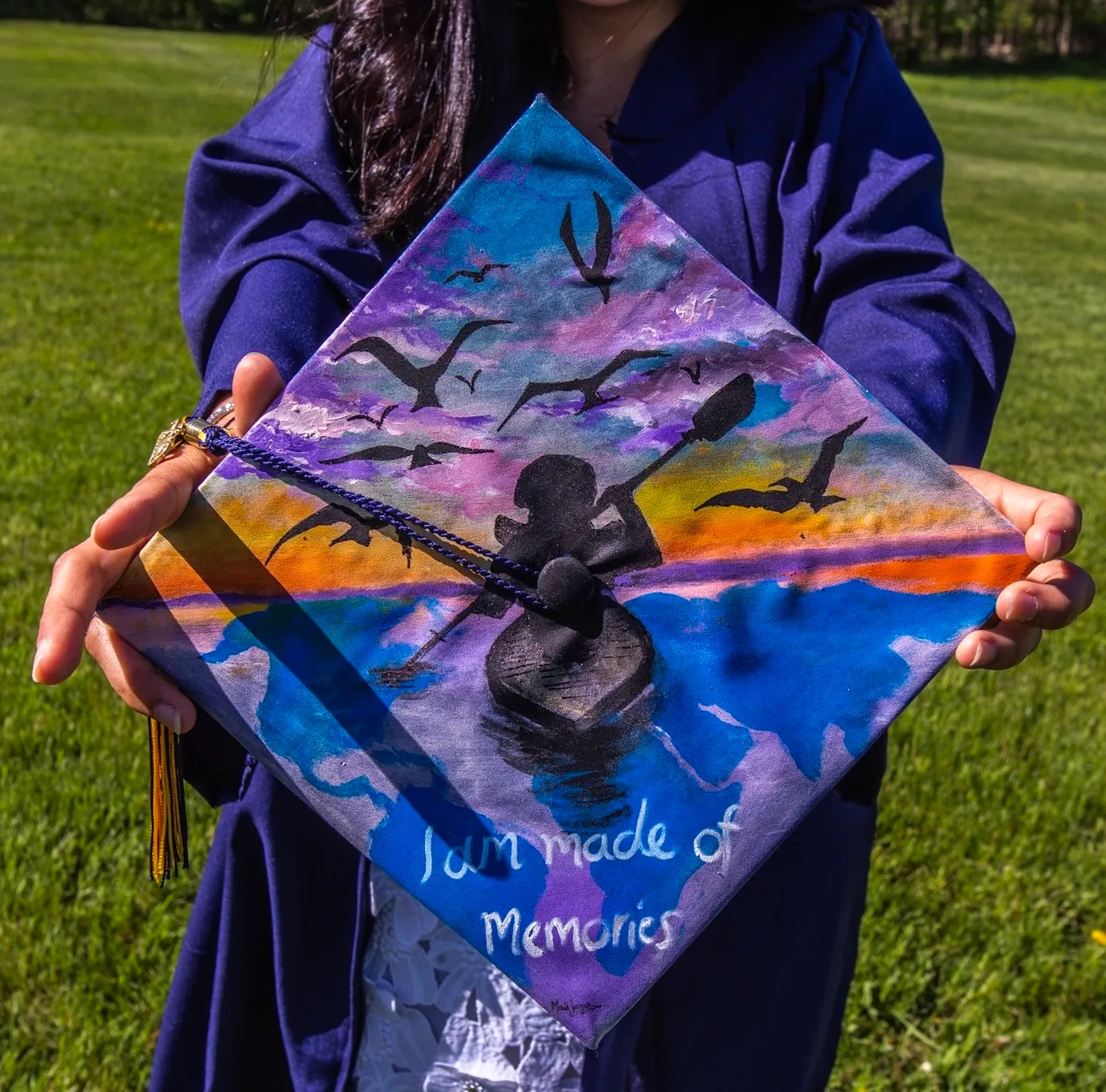 Close-up of a graduation cap held by a person in a navy blue gown, featuring a painted design of a person kayaking at sunset with flying birds and the words 'I am made of memories'.
