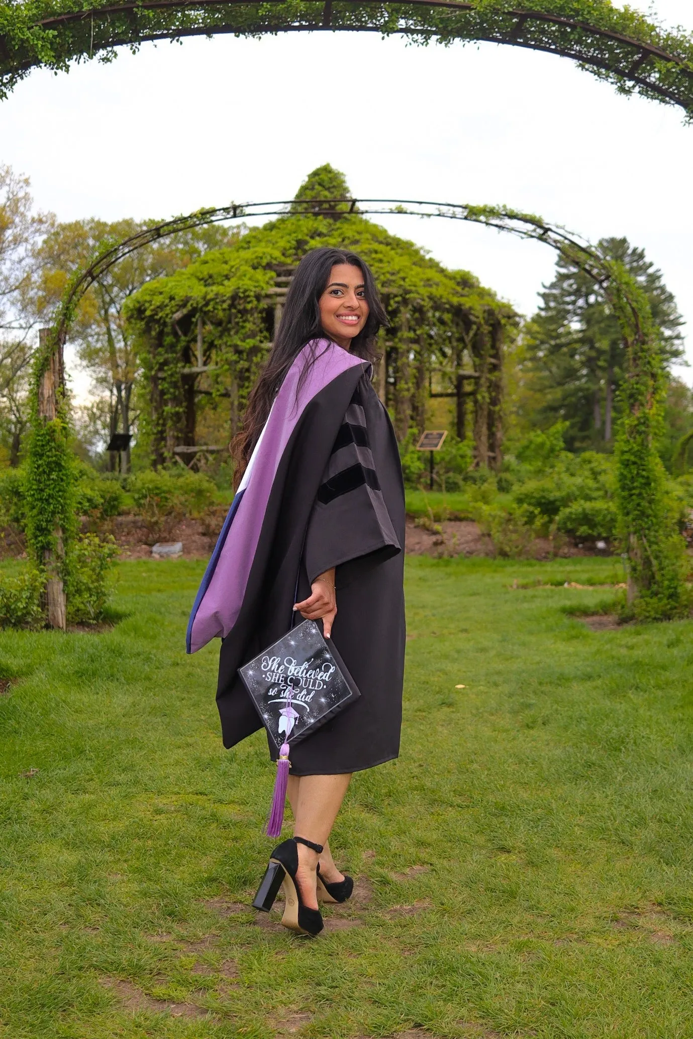 Smiling woman in graduation gown and high heels holding decorated cap saying 'She believed she could, so she did' in a green garden.