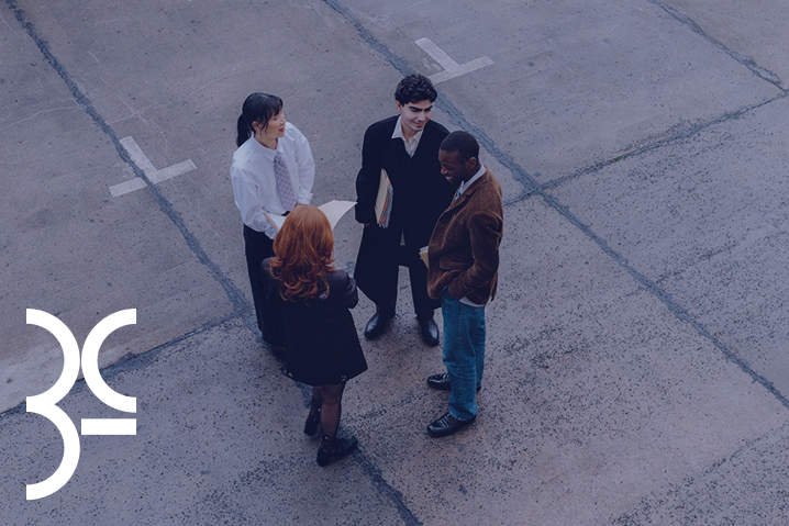 Four professionals standing in a circle talking on a concrete pavement area, holding folders and papers.