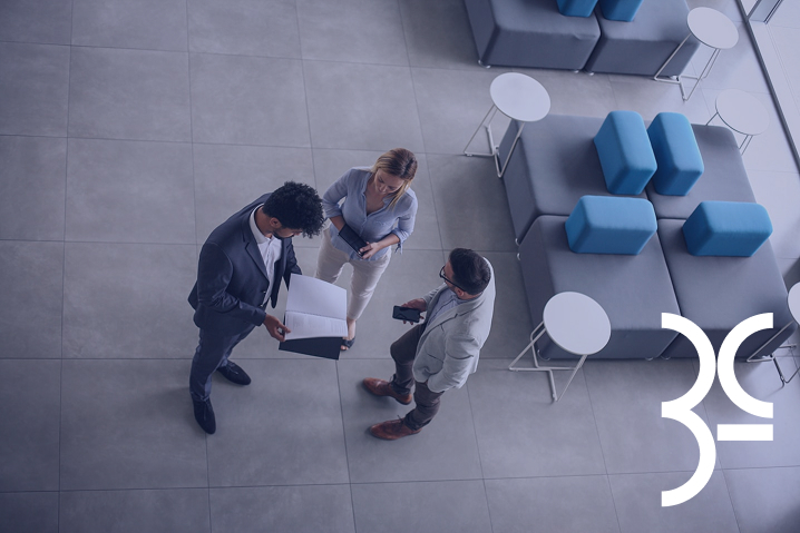Three professionals standing and discussing documents in a modern lounge area with seating and small tables.