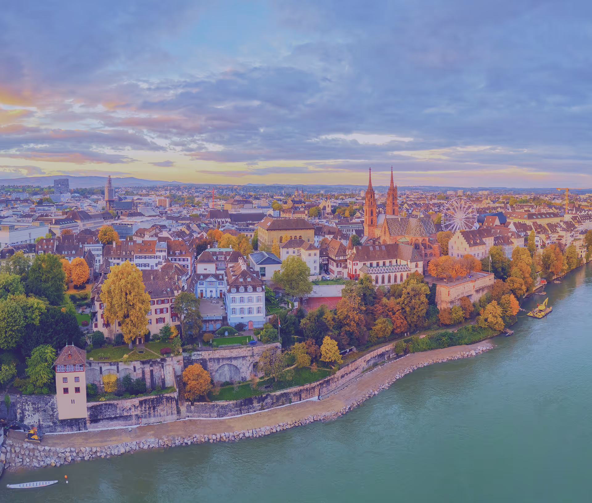 Aerial view of a European city with historic buildings, a river along the promenade, autumn trees, and a church with twin spires under a partly cloudy sky.