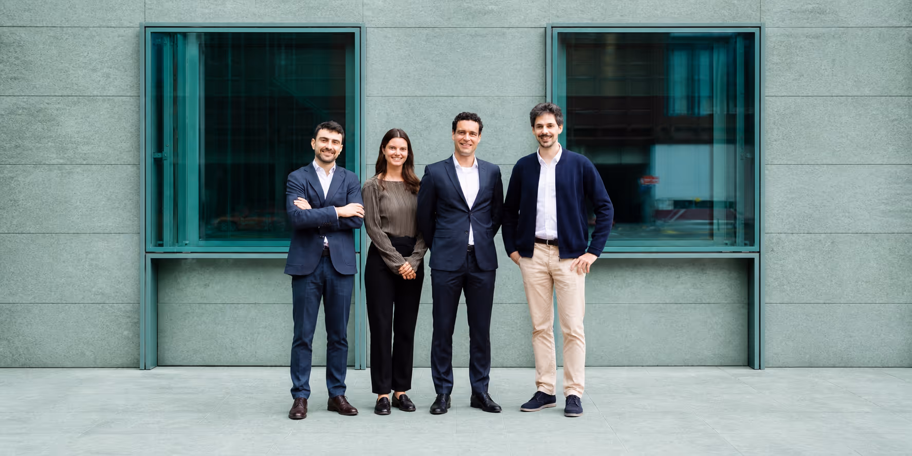 Four professionally dressed colleagues standing side by side in front of a modern building with greenish windows.