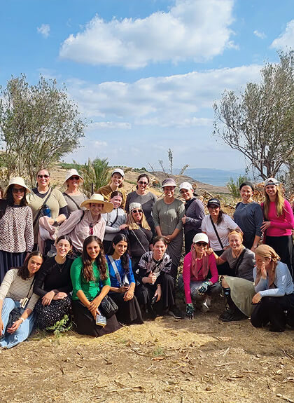 Group of women posing outdoors on a sunny day with trees and open landscape in the background.