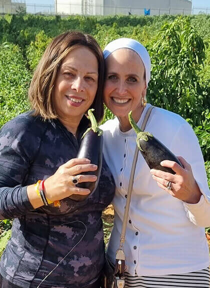 Two smiling women holding large eggplants in a lush green garden or farm field.