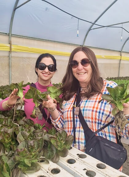 Two women smiling inside greenhouse holding green leafy vegetables with roots in a hydroponic setup.