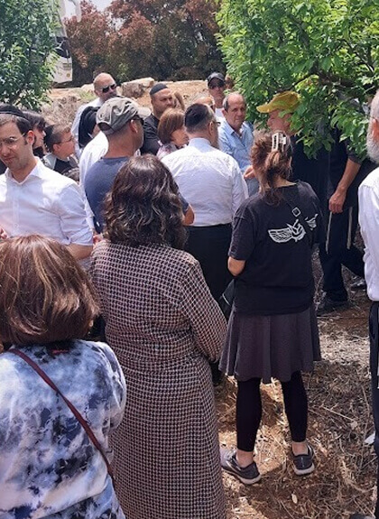 Group of people gathered outdoors near trees and bushes, some facing away and others chatting.