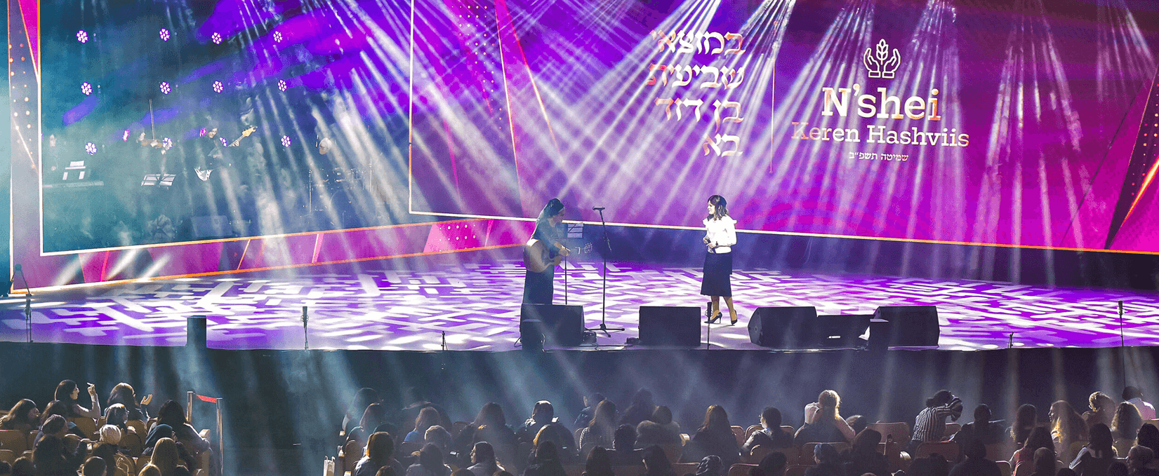 Two women on stage under colorful lights with audience watching in front at a live event.