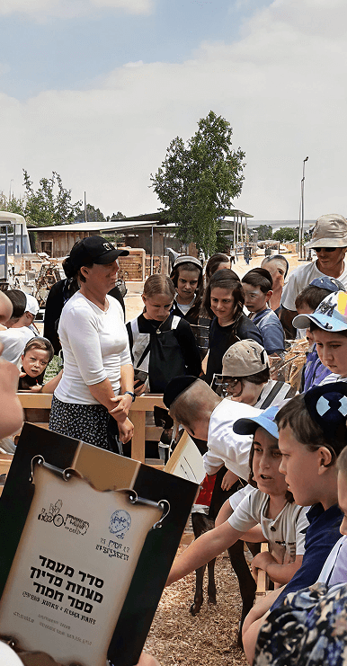 Group of children and adults gathered outdoors around a signboard in a farm setting.