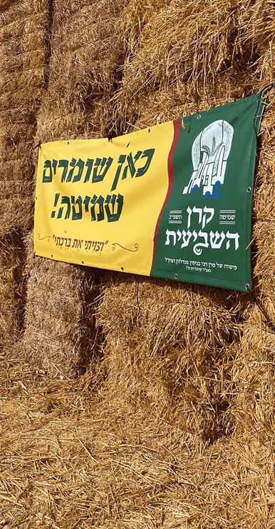 Banner in Hebrew about grain storage hung on stacked hay bales in a farm setting.