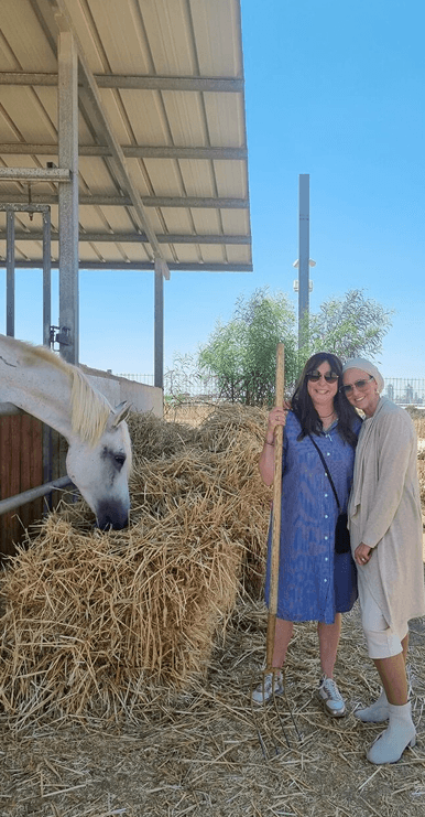 Two women smiling beside a horse eating hay under a metal roof on a sunny day.