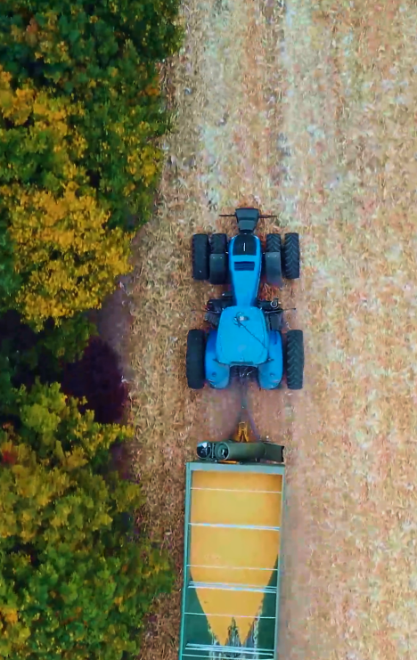 A blue tractor pulling a trailer in a field next to a dense green and yellow forest area.