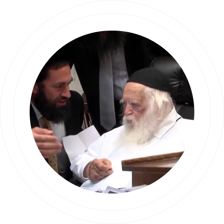 Older man with white beard and black hat sitting and talking to younger man with beard and suit.
