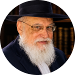 Elderly man with a white beard wearing glasses and a black hat in front of bookshelves.