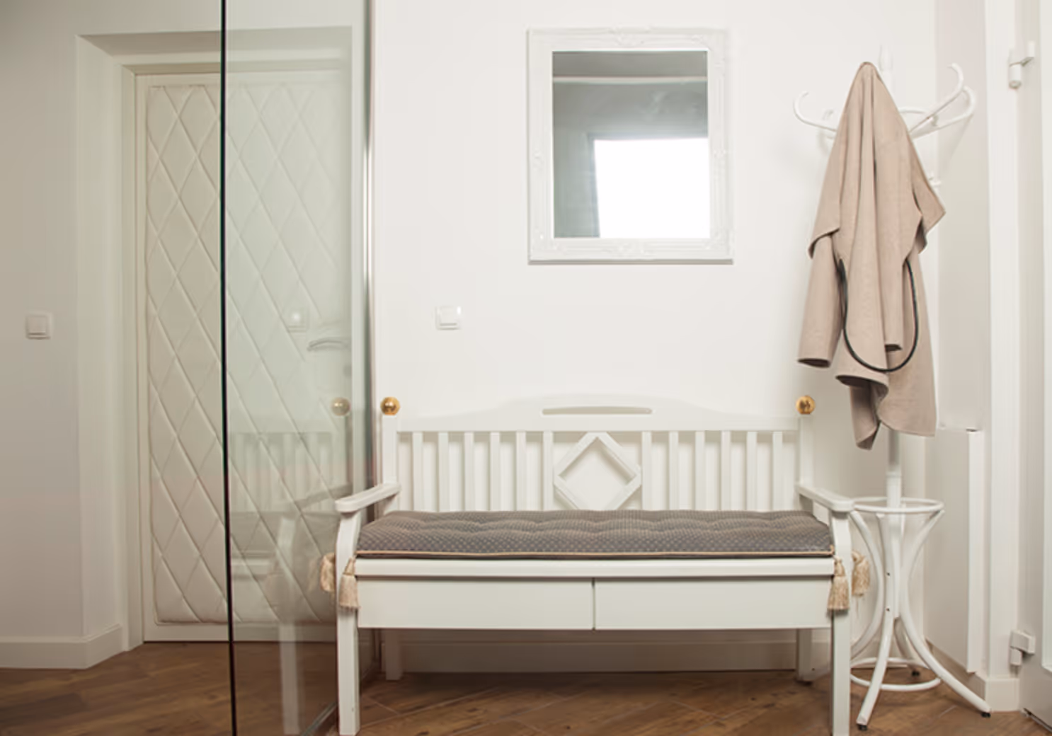 Weißer Einbaubank mit grauem Sitzkissen, Spiegel an der Wand und weißer Garderobenständer mit beigem Mantel in einem Flur.