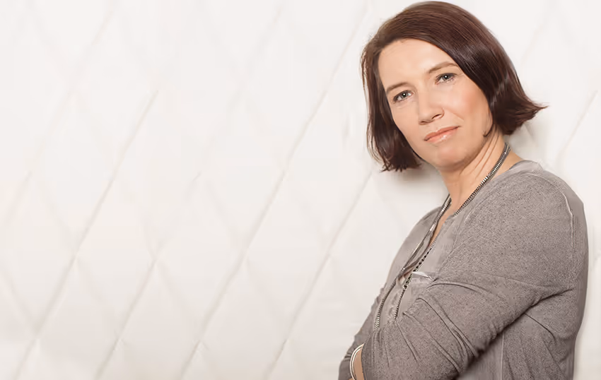 Porträt einer Frau mit kurzen braunen Haaren, die vor einem weißen gepolsterten Hintergrund steht.