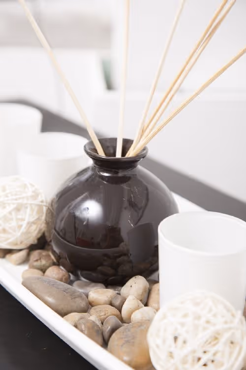Schwarze Duftöllampe mit Holzstäbchen auf weißen Steinen, umgeben von weißen Kerzenhaltern und dekorativen Kugeln.