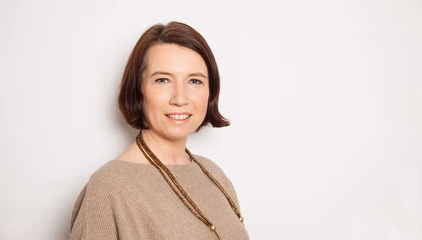 Frau mit kurzen braunen Haaren trägt einen beigen Pullover und eine zweireihige Perlenkette vor einem weißen Hintergrund.