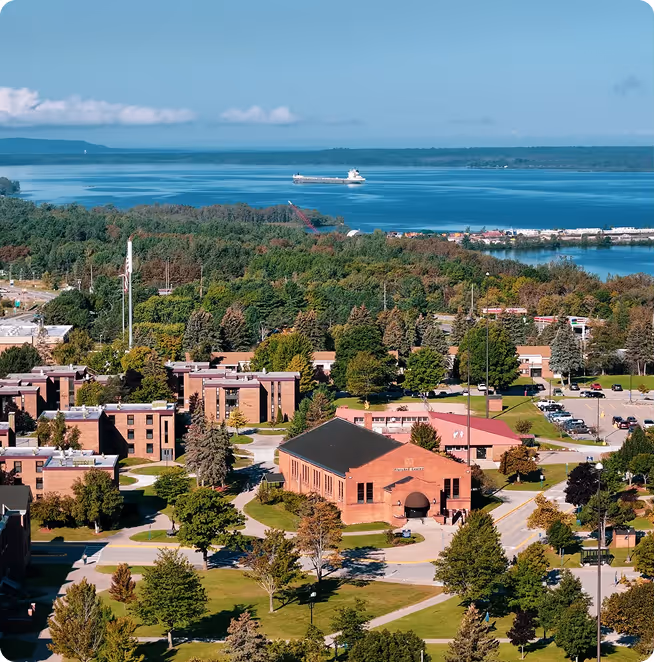 Aerial view of a campus with brick buildings surrounded by green trees, with a large body of water and a ship in the background.