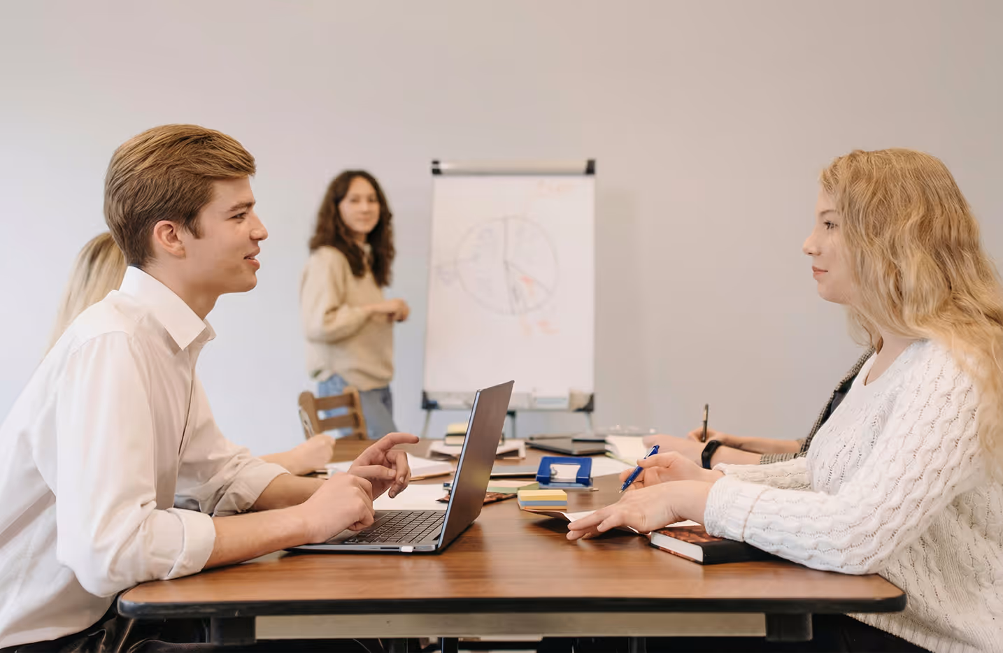 Young man and woman sitting across a table with laptops and notebooks, having a discussion in a meeting room with a presenter and a flip chart in the background.
