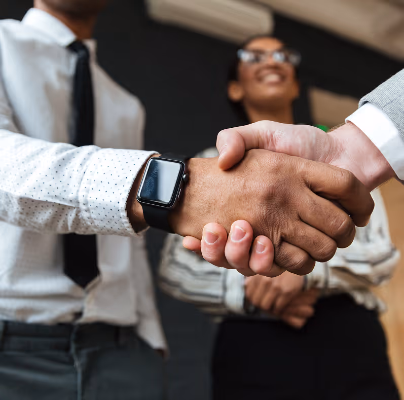 Two businesspeople shaking hands in an office, with a smiling woman blurred in the background.
