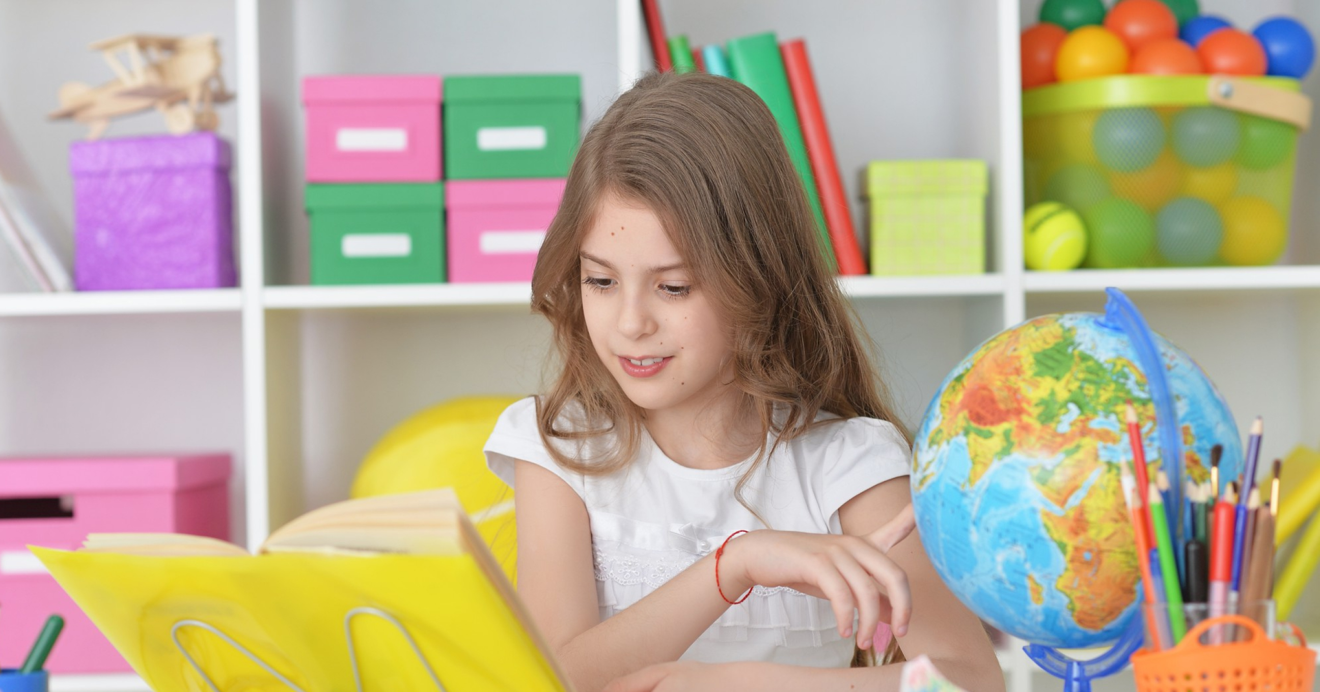 Young girl with long hair reading a book at a desk with colorful stationery and a globe nearby.