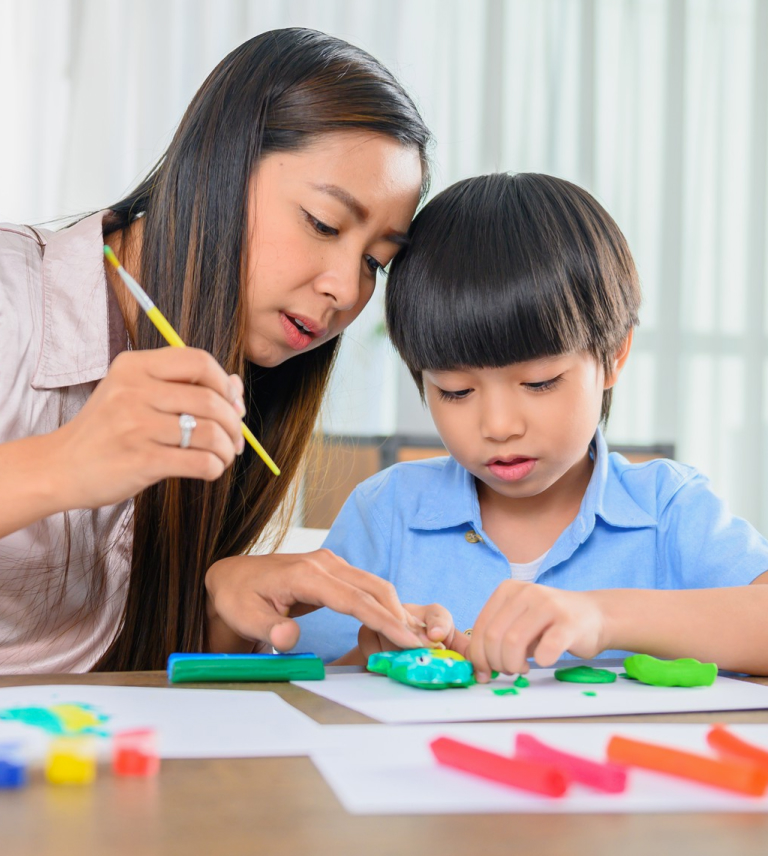 Woman closely guiding a young boy as they work together with colorful modeling clay at a table.