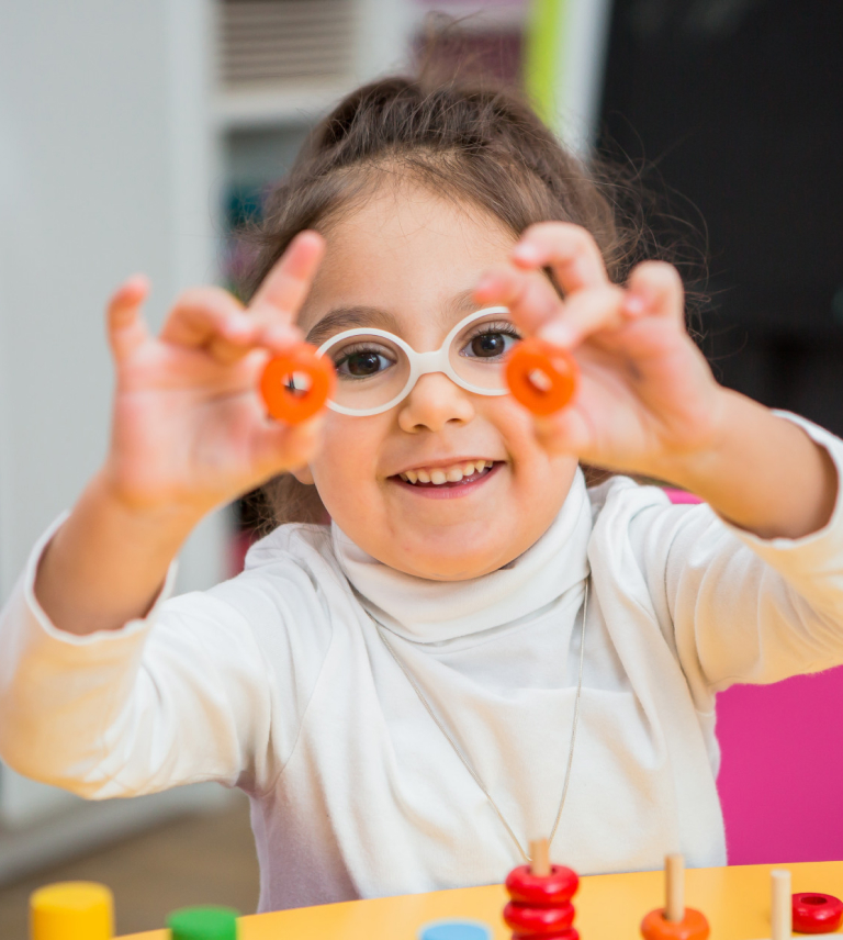 Smiling girl wearing glasses holding two orange rings in front of her face while playing with colorful wooden toys.