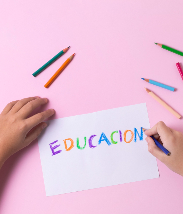 Child's hands writing the word EDUCACION in colorful letters on white paper using colored pencils on a pink background.