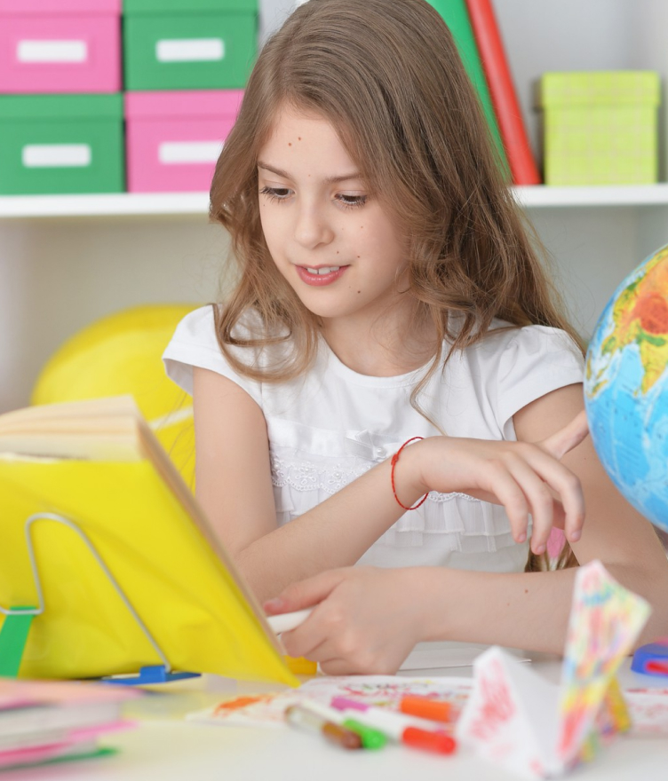 Young girl reading a book with a globe and colorful markers on a desk in a bright room.