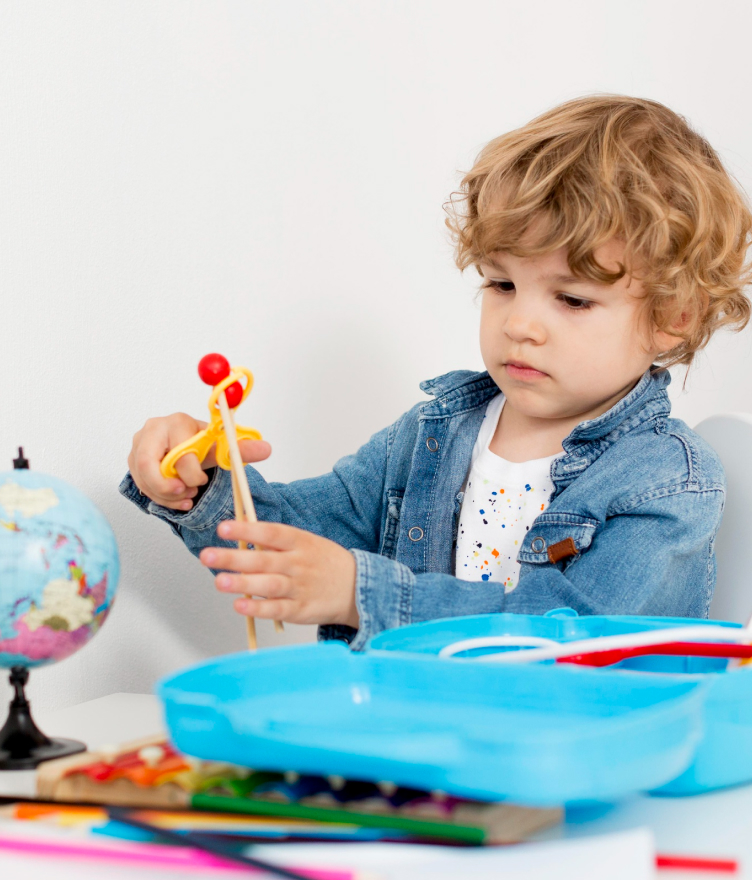 Young child with curly blonde hair in a denim jacket playing with a toy bow and arrow at a table with art supplies and a globe.