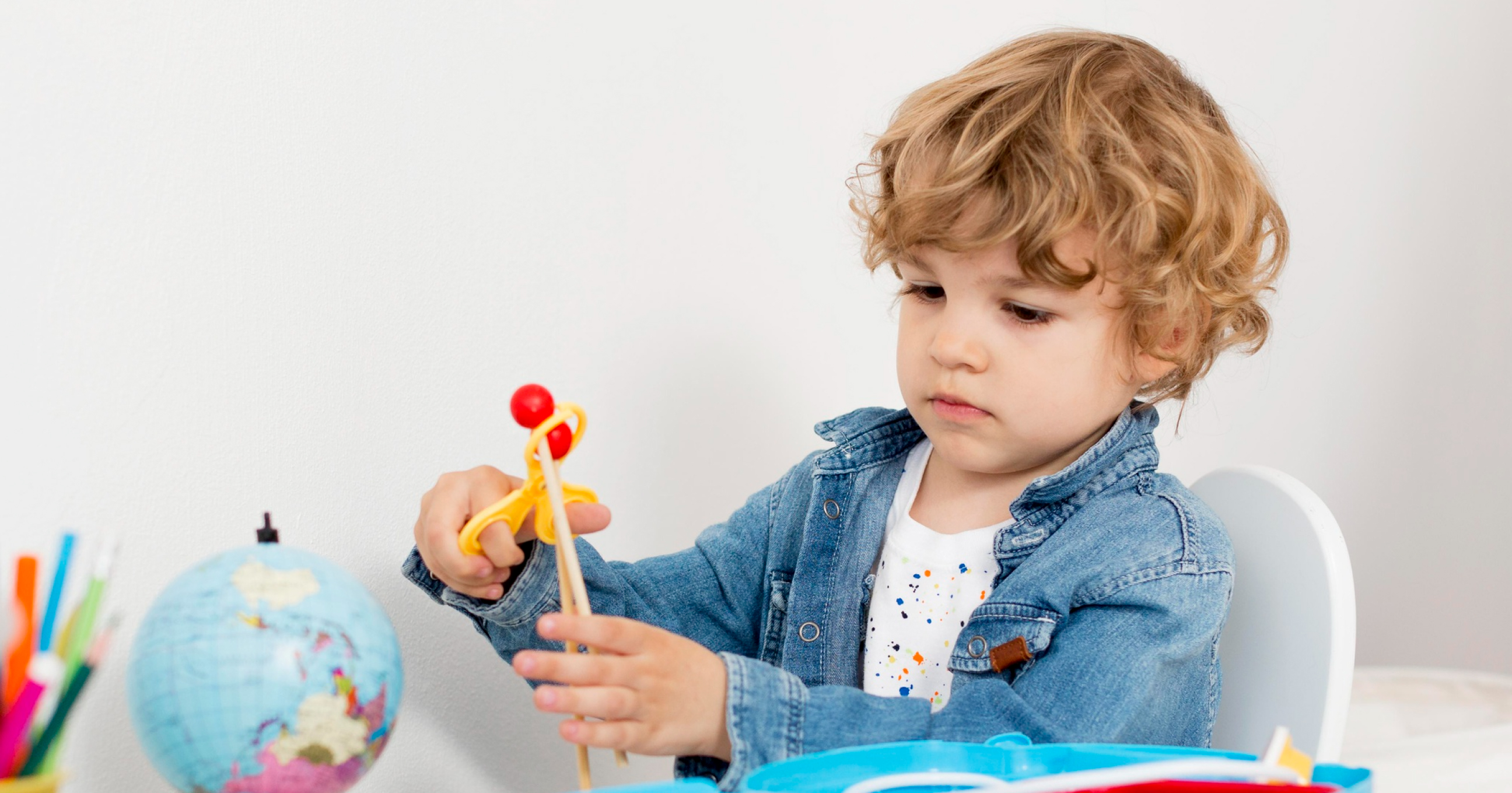 Young boy with curly hair wearing a denim jacket, playing with a yellow toy slingshot at a table with a globe and colorful pencils.