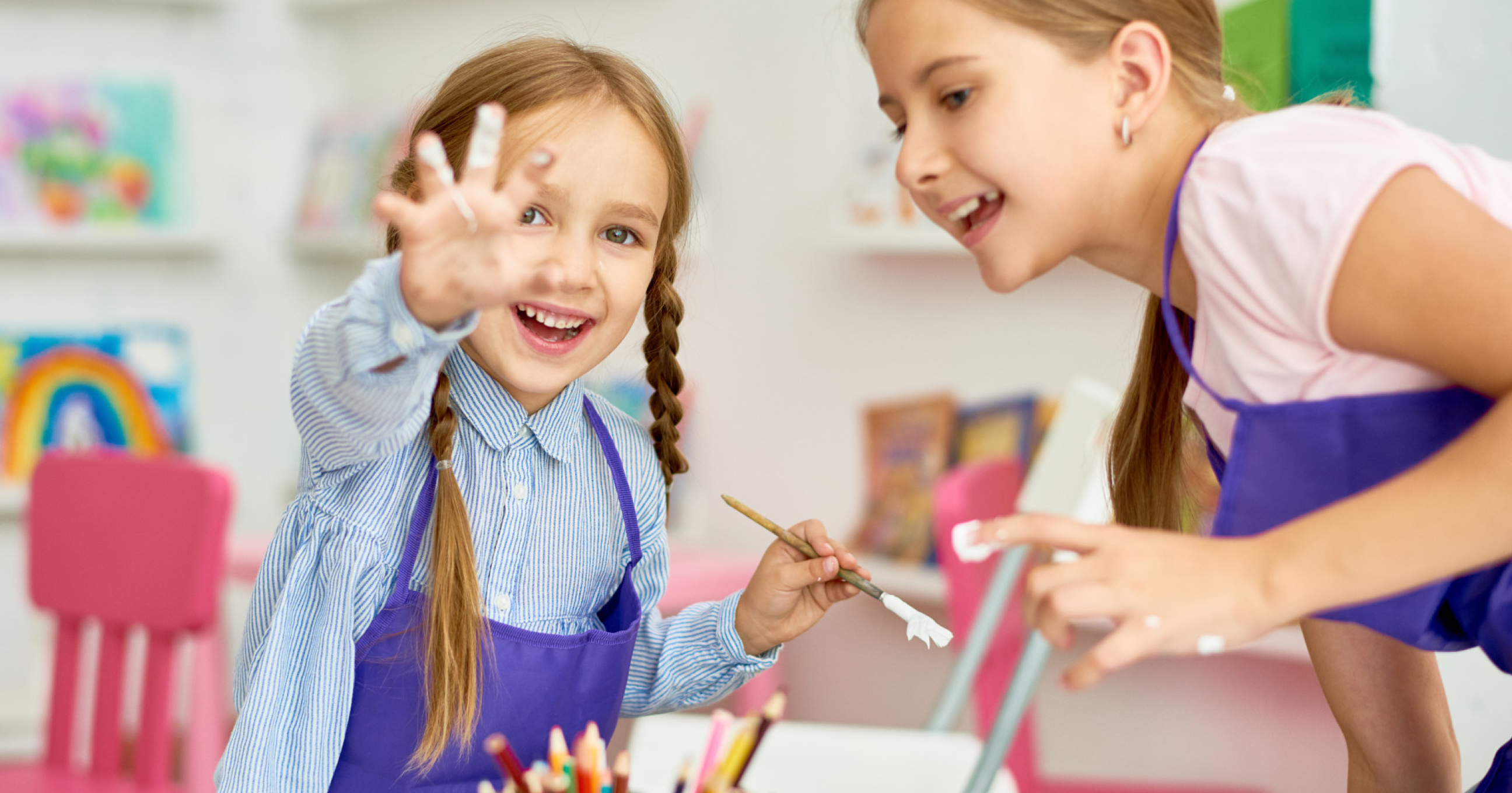 Two young girls wearing purple aprons are painting and smiling, one showing her paint-covered fingers.